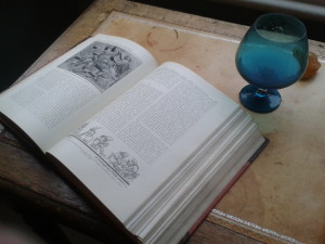 A book containing Roman bas relief pictures on an leather covered desk and a blue brandy glass next to it